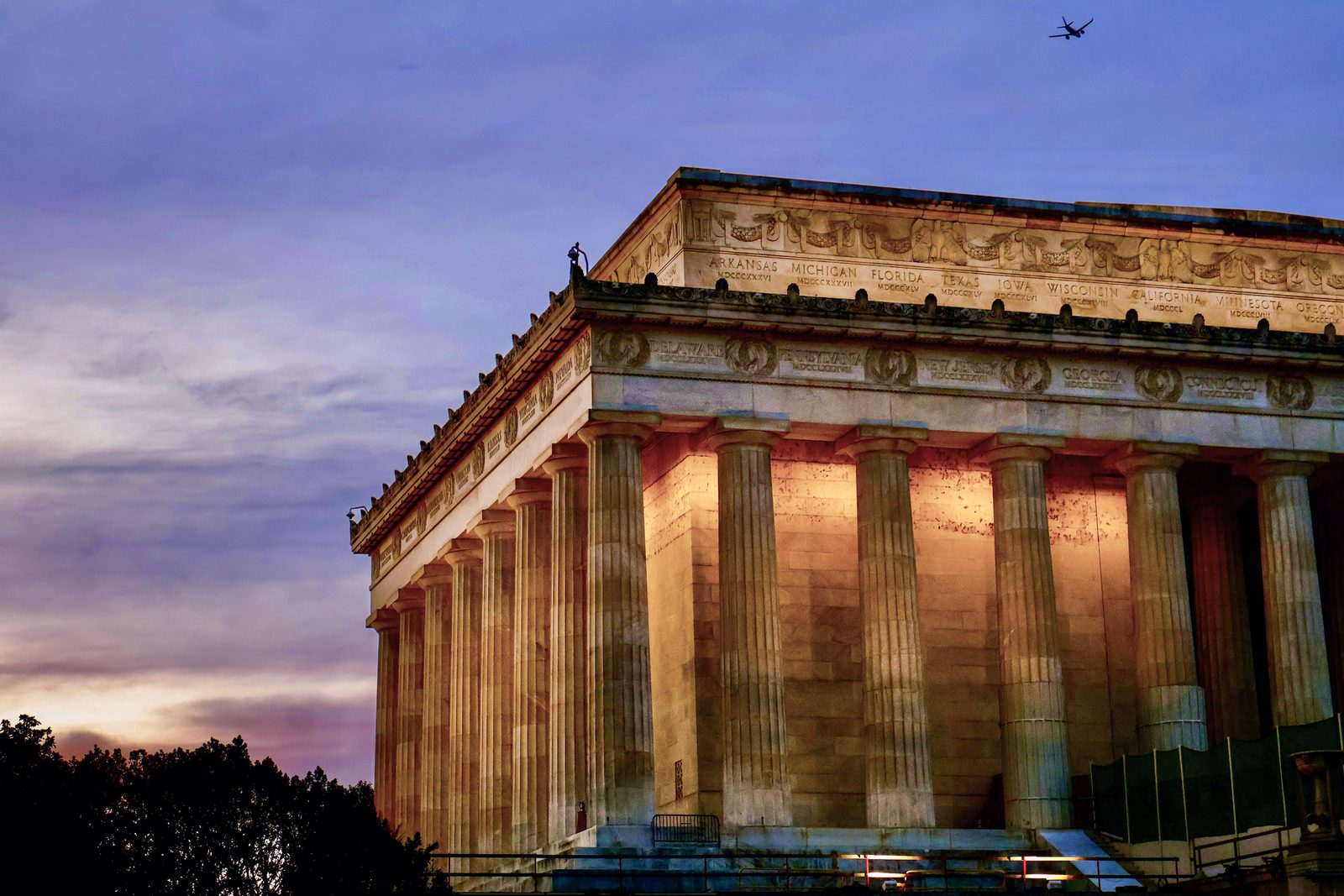 A low-angle view of the Lincoln Memorial at last light, a plane crossing a deep mauve sky.