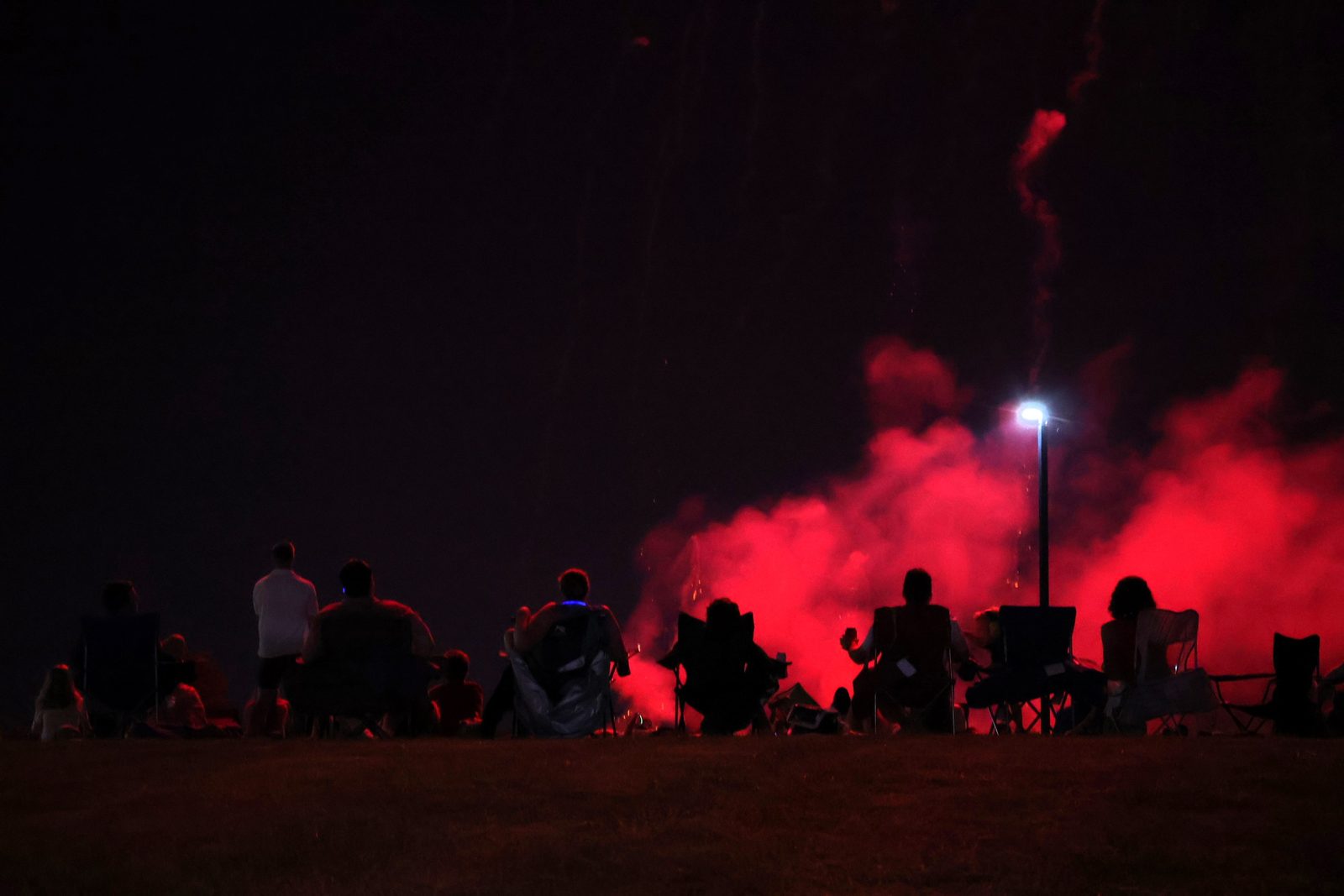 A crowd in lawn chairs at night, silhouetted against a wash of red flare smoke and a single bright stadium lamp.
