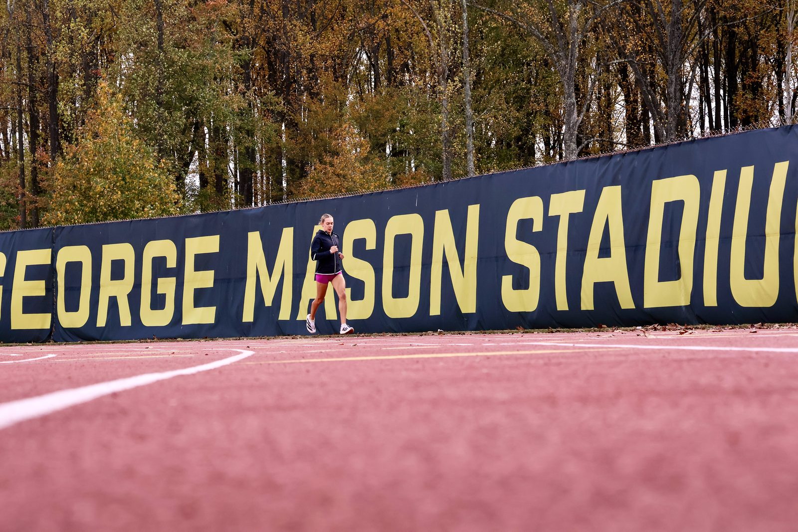 A solitary cross-country runner passing a navy and gold George Mason Stadium banner.