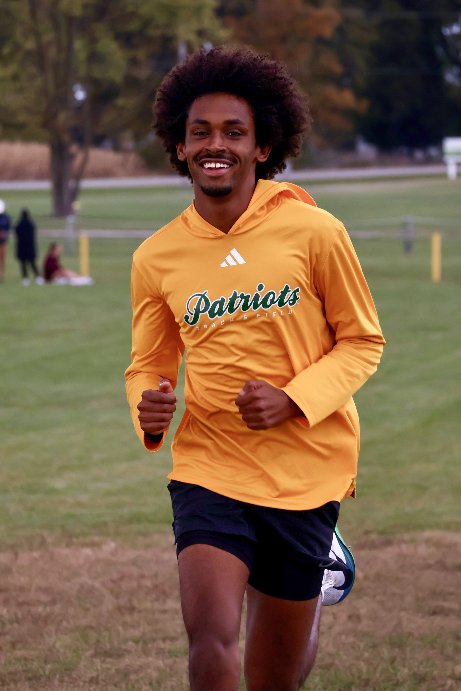 George Mason 'Patriots Track & Field' runner smiling mid-stride in an open field.
