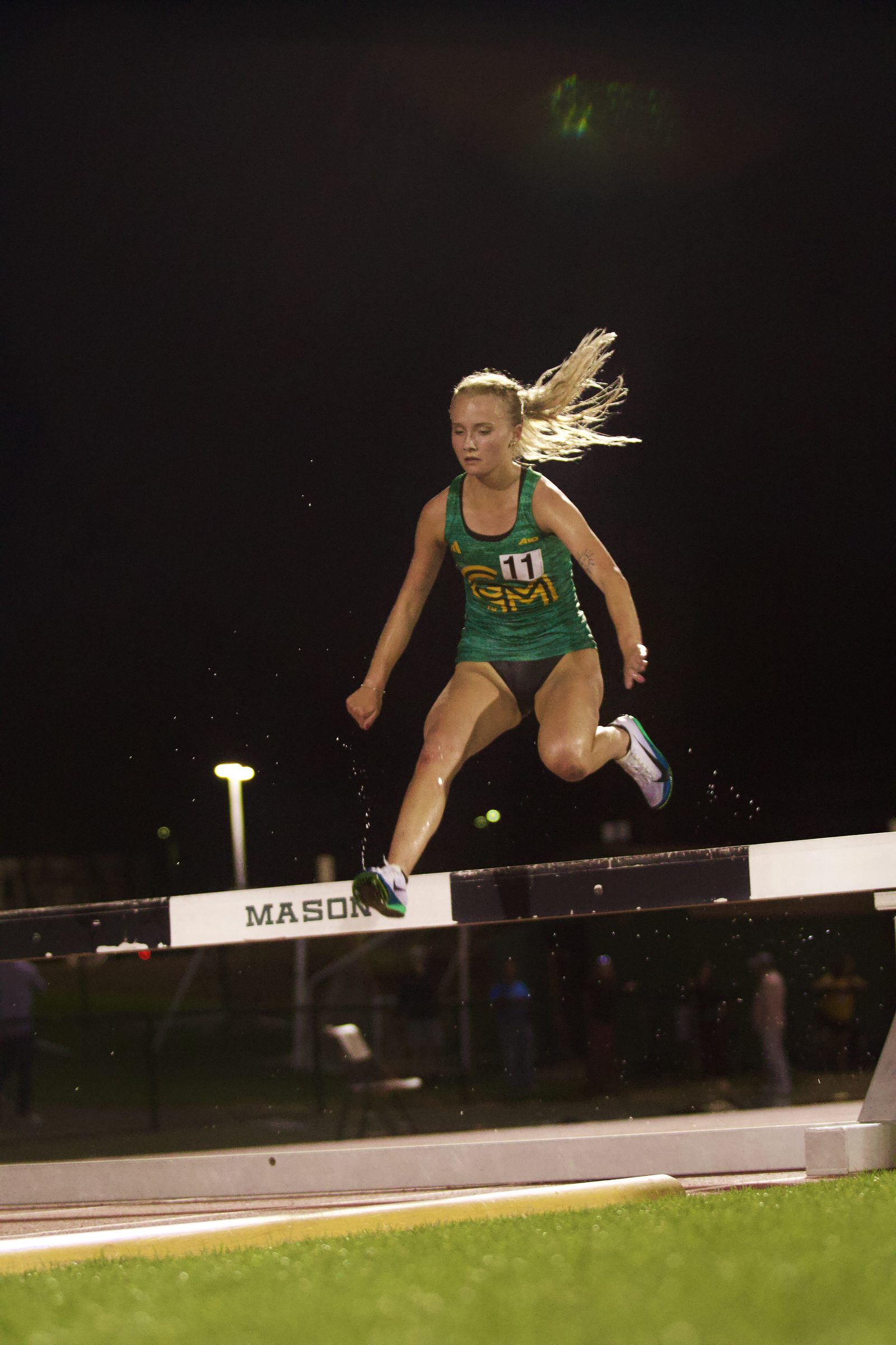 Steeplechase runner clearing a water barrier at a night meet, hair flying behind her.