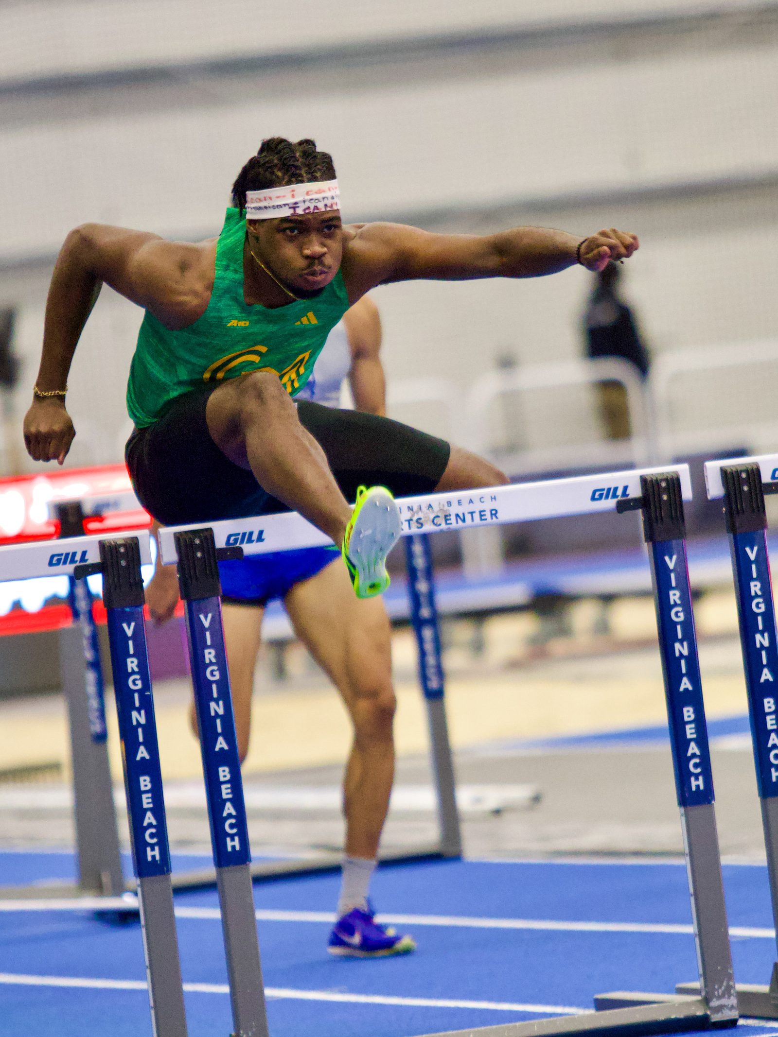 Sprint-hurdler mid-clearance over a hurdle at an indoor track meet.