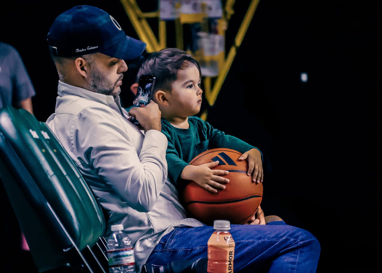 A father holds a toddler in the arena seats; the child clutches a basketball, watching the court.