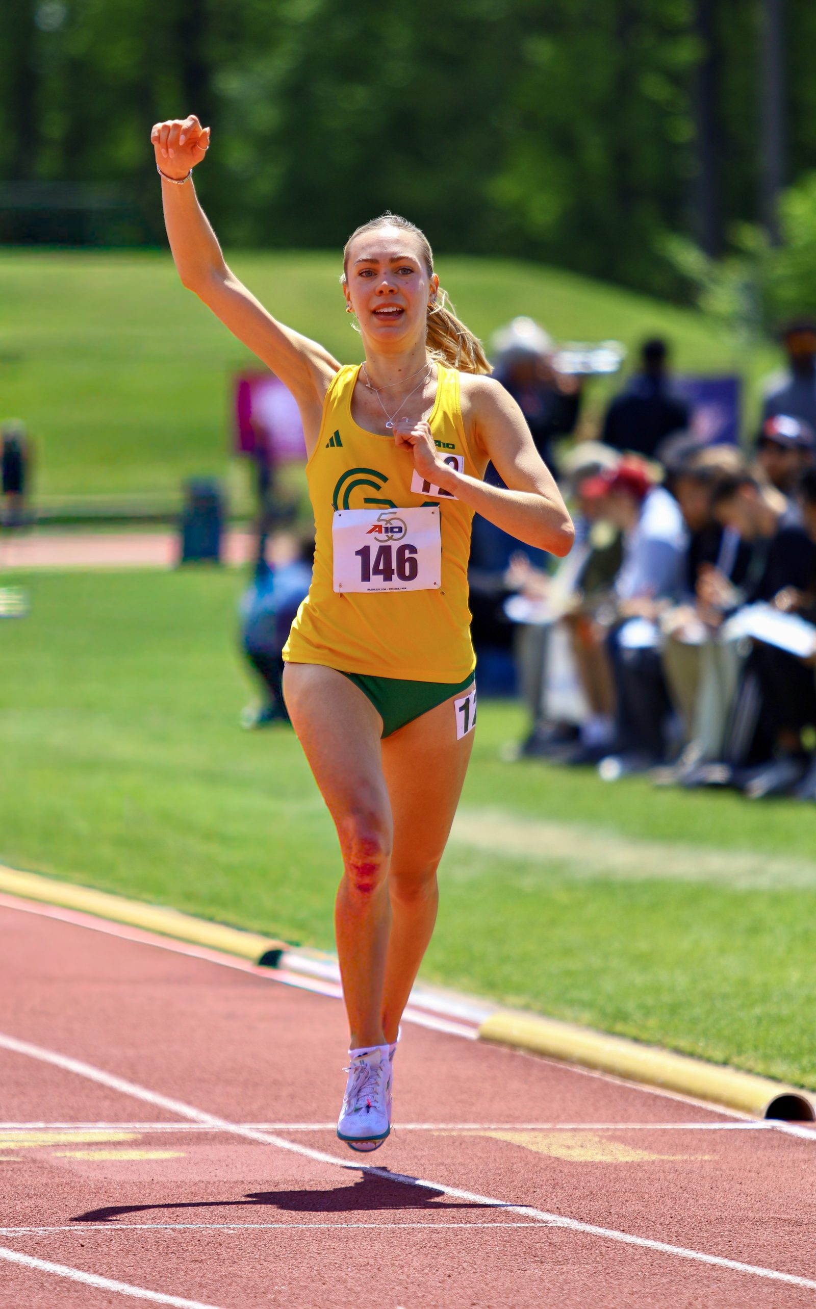 George Mason runner in a yellow singlet, bib 146, crossing the finish line with fist raised in celebration on a sunny day.
