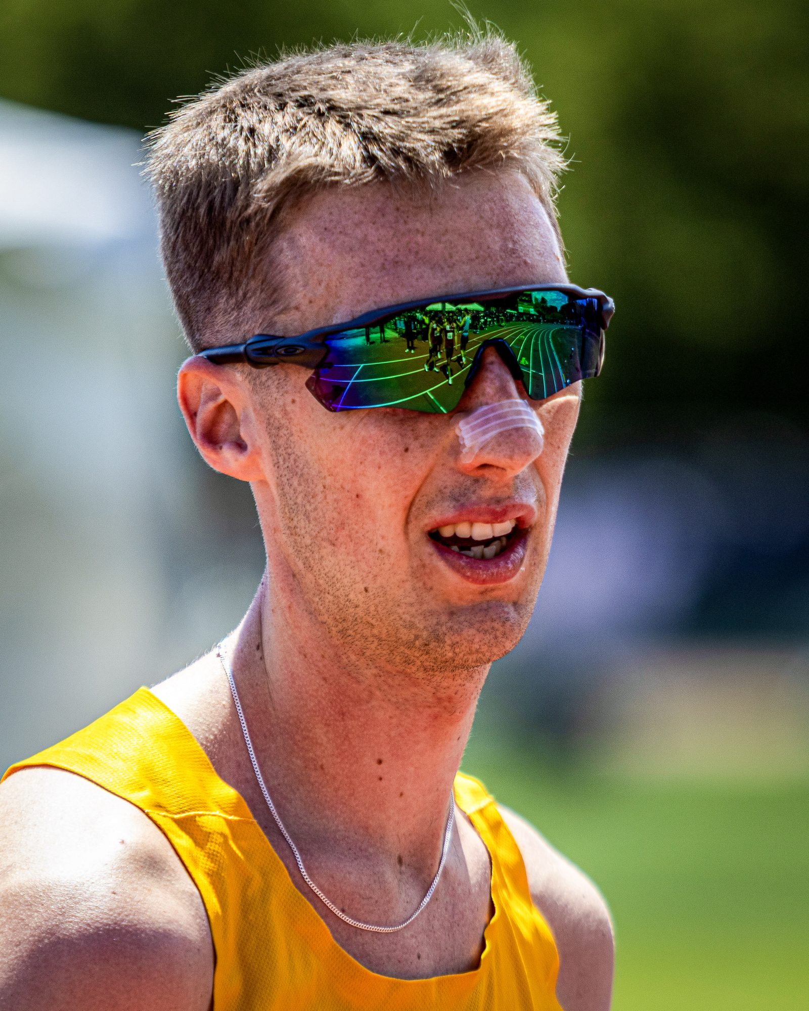Tight portrait of a George Mason runner wearing mirrored sport sunglasses that reflect the infield, nose strip on his face.