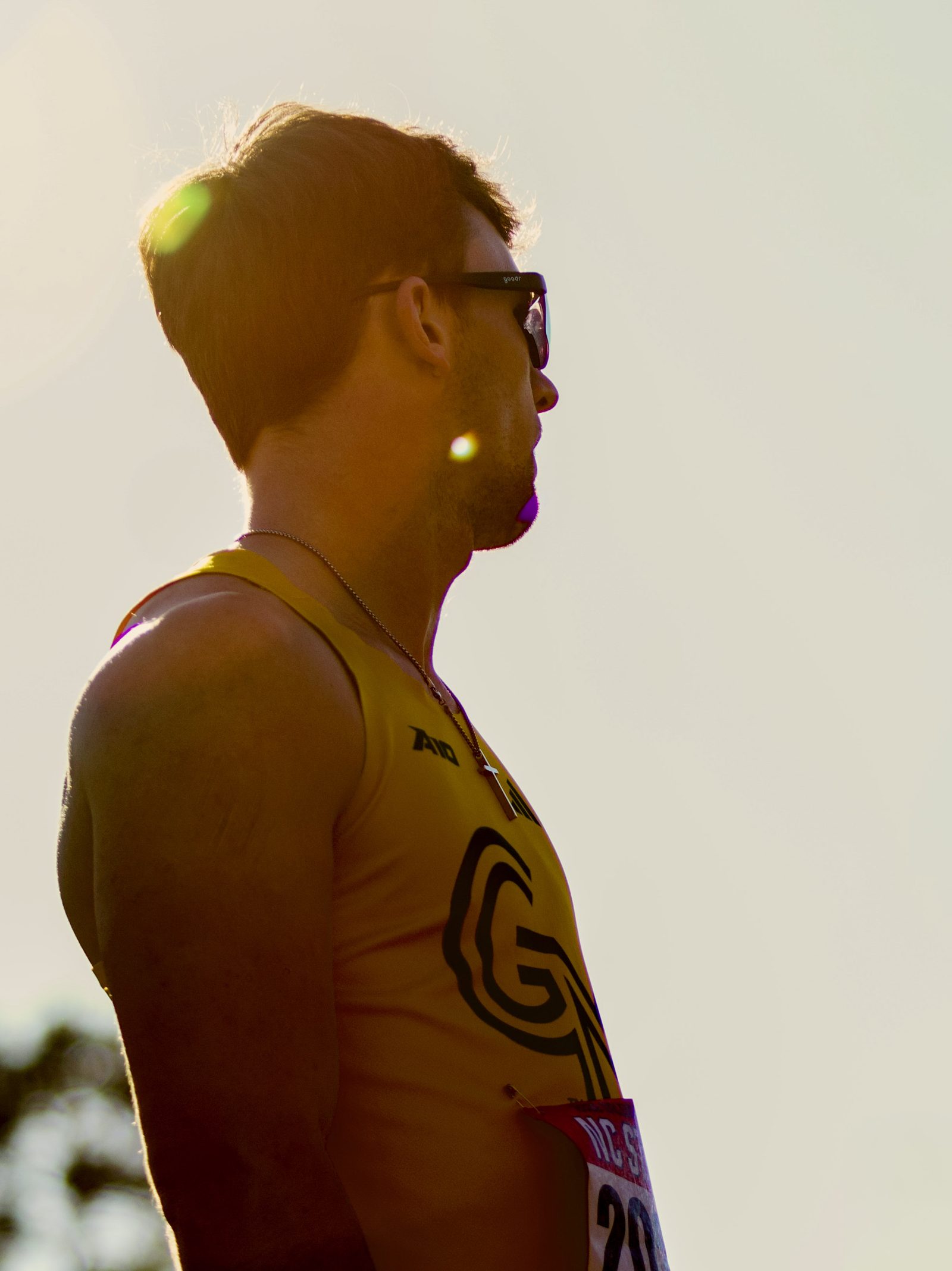 Backlit profile portrait of a George Mason athlete in a yellow singlet, late-day sun flaring across the frame.