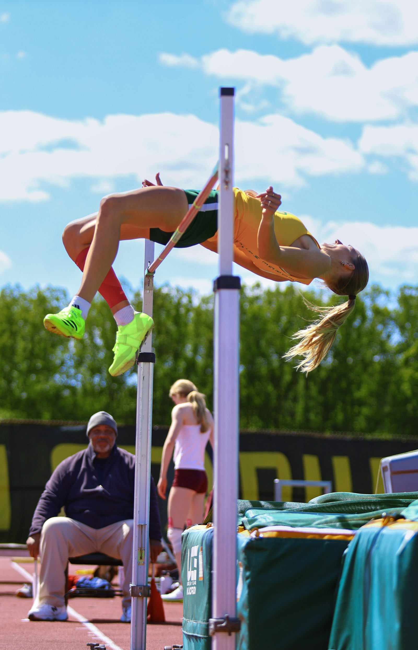 Female high jumper arching backward over the bar against a blue sky, an official watching from below.