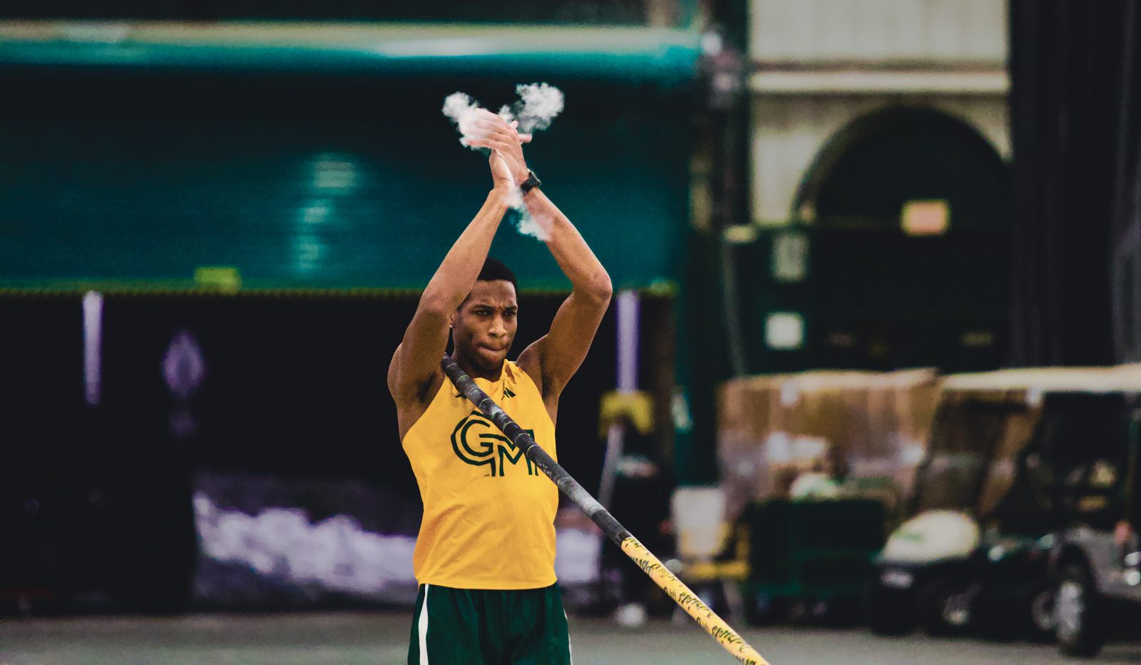 George Mason pole vaulter clapping chalk dust between his hands before a jump, indoor track facility in the background.