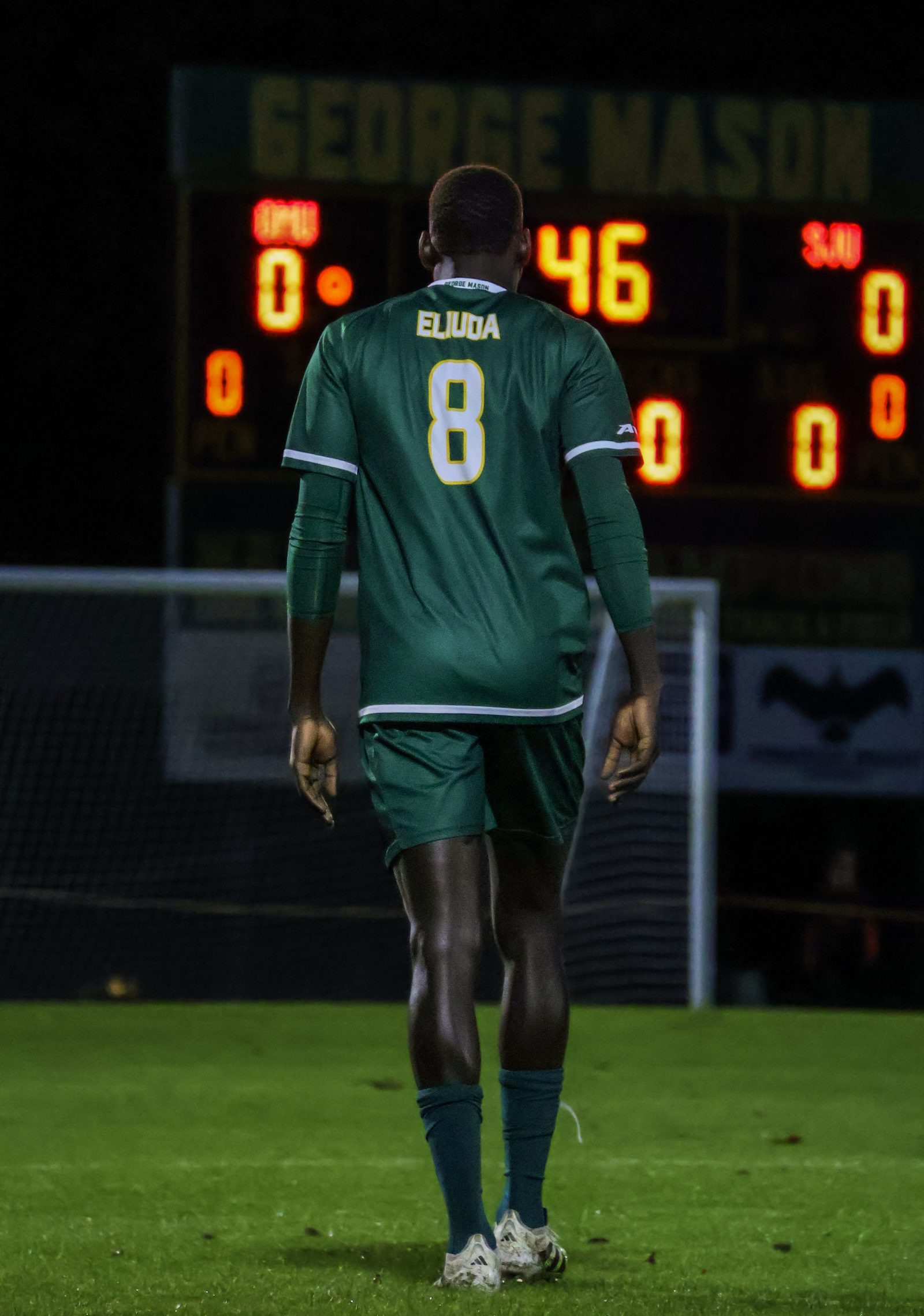 George Mason soccer player "Eljuda," number 8, photographed from behind walking on the field, with the stadium scoreboard glowing behind him.